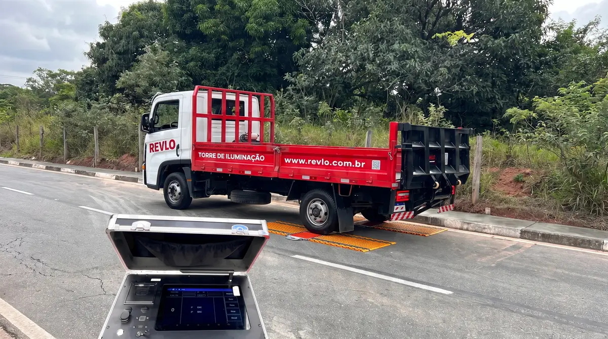 Caminhão Revlo de carroceria vermelha com a roda traseira sobre as plataformas laranja da balança móvel, numa rua asfaltada, com a maleta do display aberta em primeiro plano