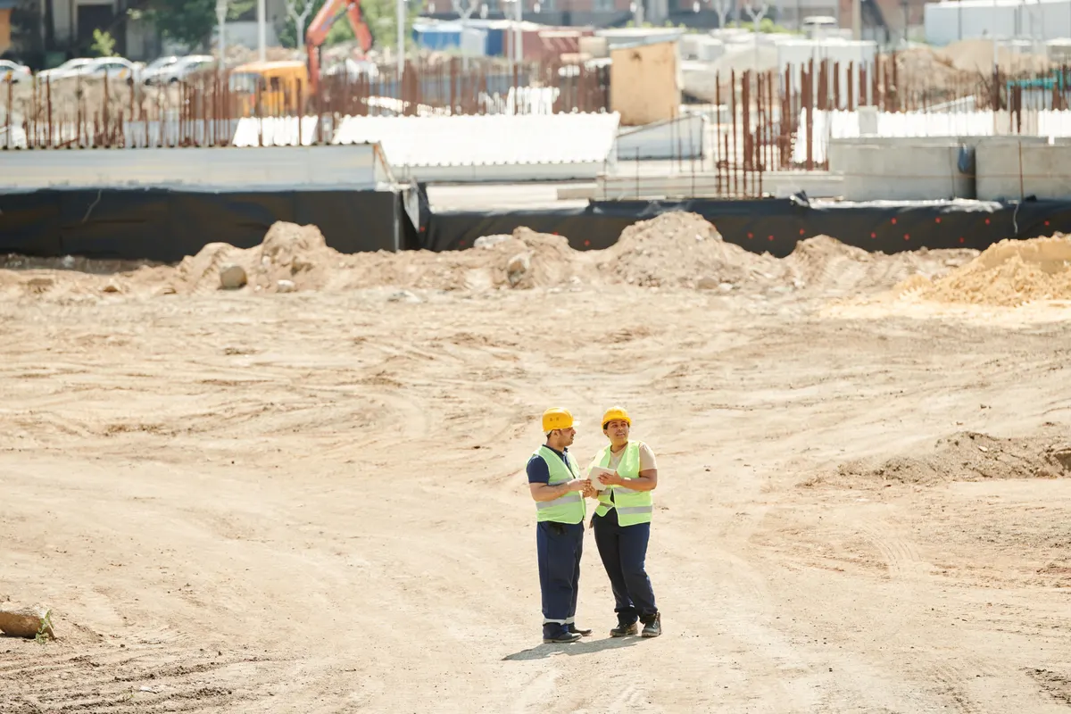 Dois trabalhadores de colete conversando em área ampla de terraplanagem