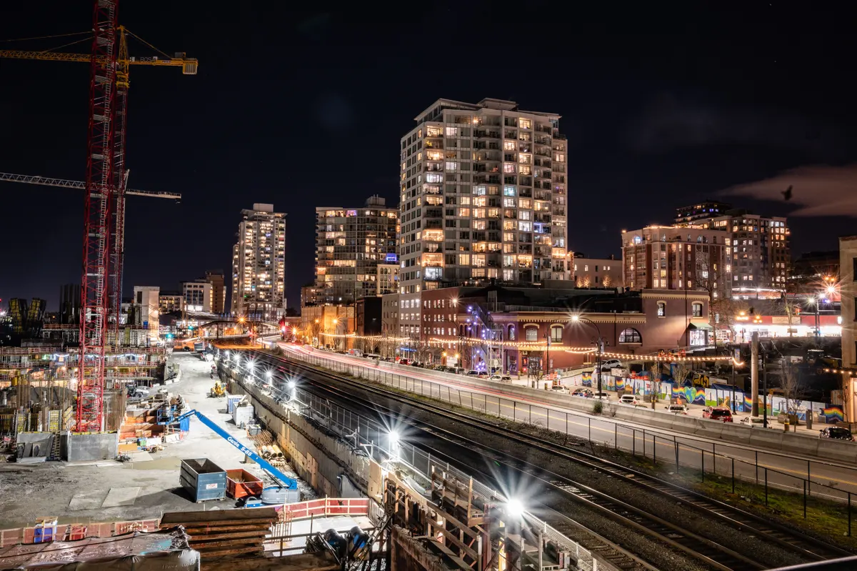 Canteiro de obras urbano com guindastes e prédios residenciais ao fundo durante a noite