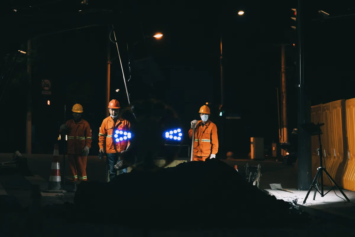 Dois trabalhadores de colete refletivo em frente de serviço noturna iluminada por refletor portátil