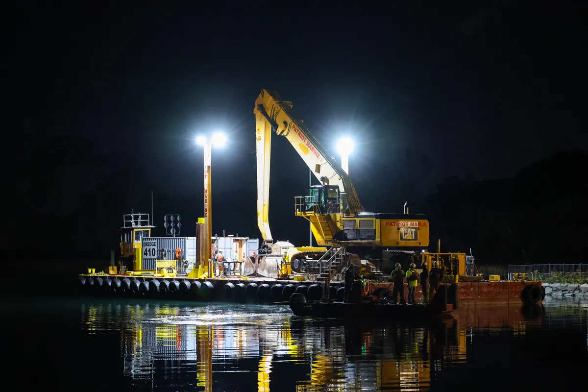 Escavadeira sobre balsa operando à noite sob refletores, com o reflexo na água