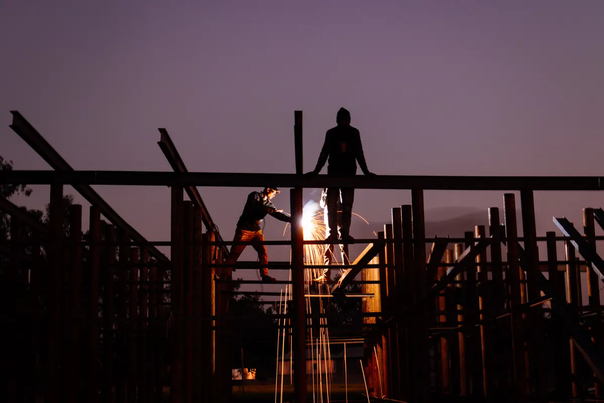 Silhueta de trabalhadores sobre estrutura metálica em construção no fim da tarde