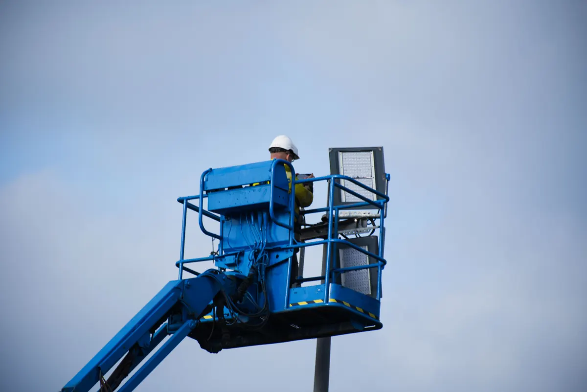Operário de capacete em cesto de plataforma elevatória azul, visto de baixo contra o céu
