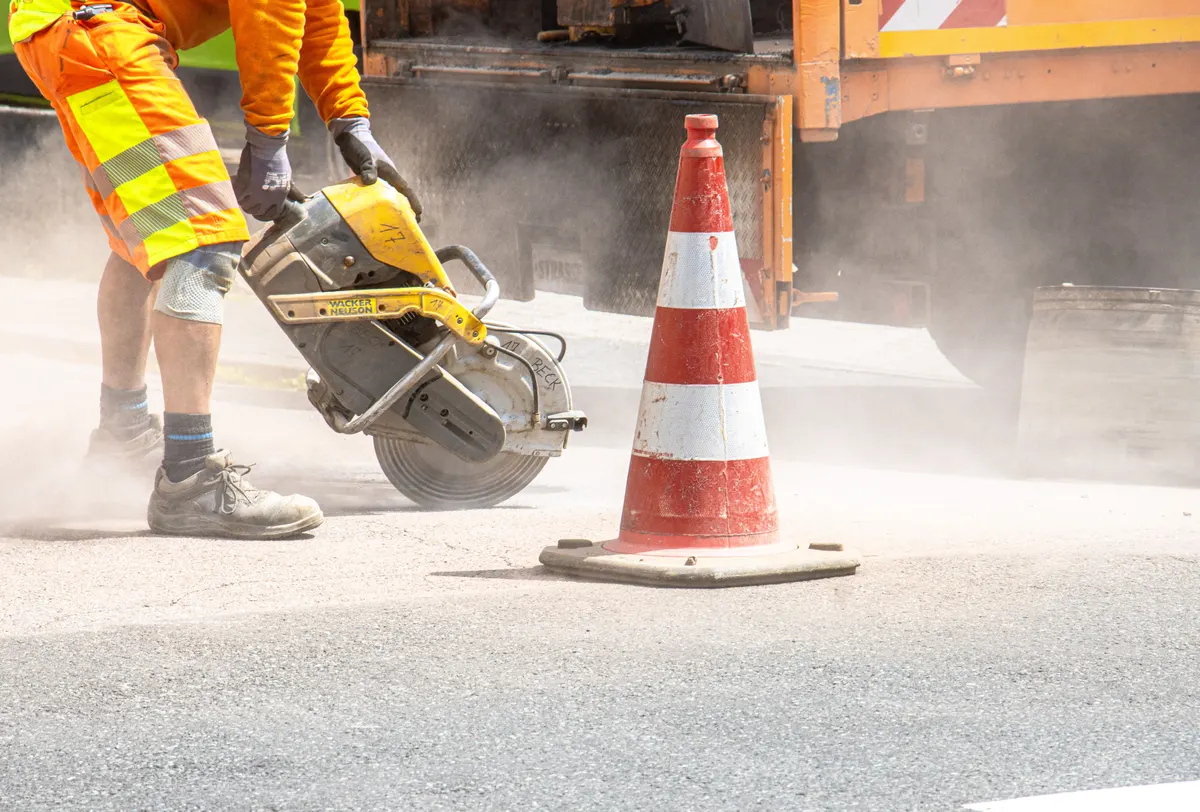 Operário de colete cortando asfalto com serra, ao lado de cone de sinalização