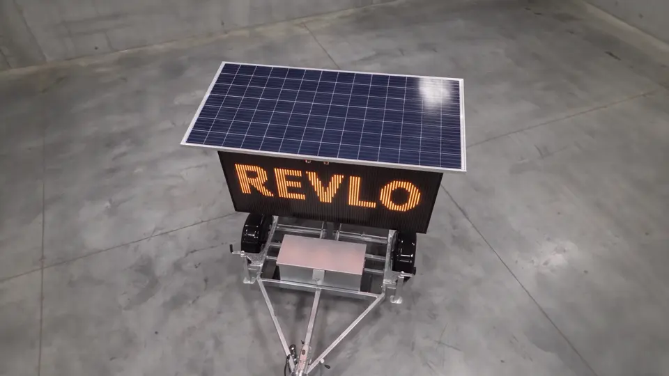 Painel de mensagem variável móvel Revlo dentro do galpão, visto de cima em ângulo, com a palavra REVLO acesa em LED âmbar e a placa fotovoltaica aberta sobre a tela